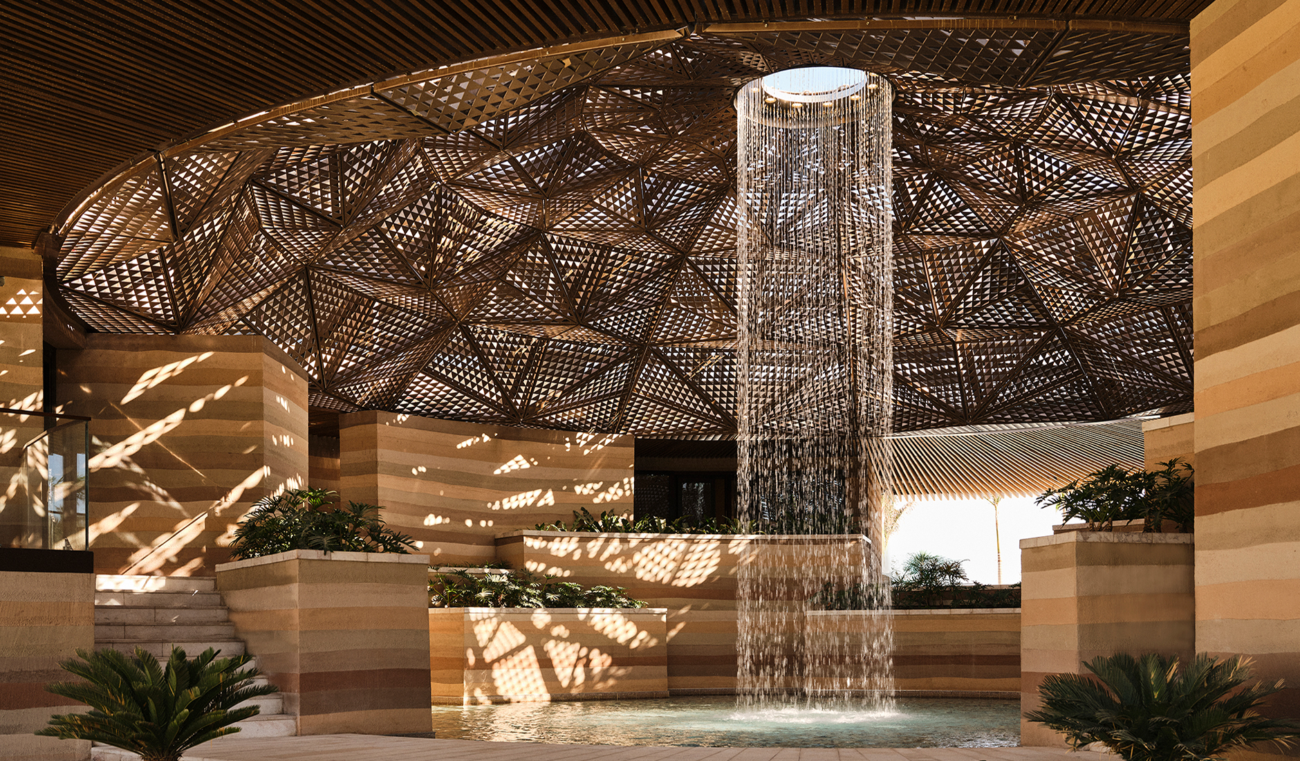 The extraordinary Hyliaa Spa arrival pavilion at Four Seasons Resort AMAALA at Triple Bay, ranked among the best luxury resorts in Saudi Arabia, featuring a majestic cylindrical waterfall cascading from a gold-toned ceiling oculus.