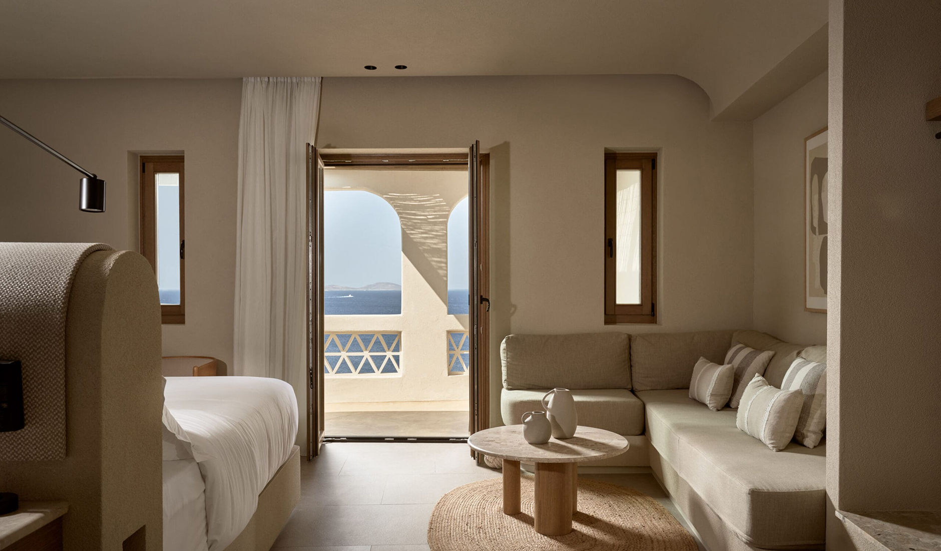 Sleek minimalist bedroom interior inside Omeon Mykonos, showcasing elegant earth-toned boho decor and large windows facing the ocean.